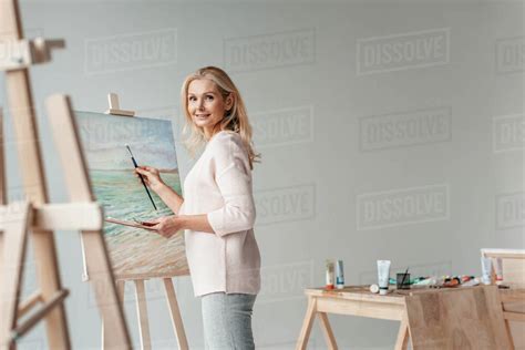 Mature Female Artist Smiling At Camera While Painting On Easel In Art