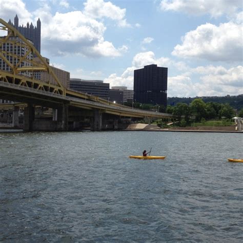 Two People In Kayaks Paddling Under A Bridge Over A River With Tall