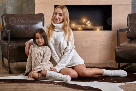 Mom with little girl relaxing on floor near a cozy sofa, Christmas lights in the fireplace ...