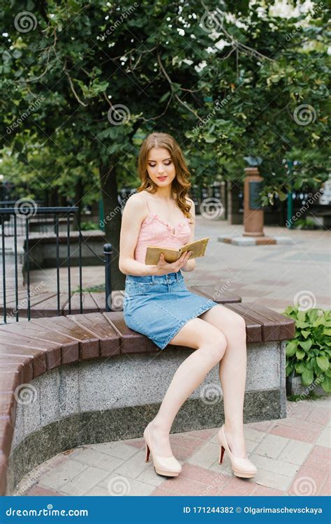 A Beautiful Romantic Girl In A Pink Top And Denim Skirt Is Reading A