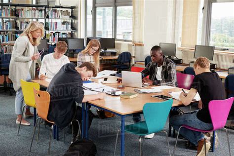 Busy College Library With Teacher Helping Students At Table Stock