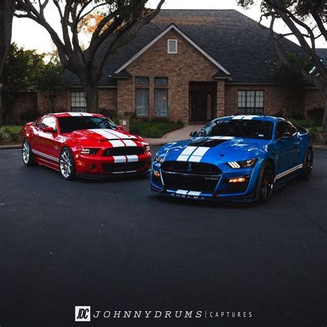 two red and blue mustangs are parked in front of a brick house with trees