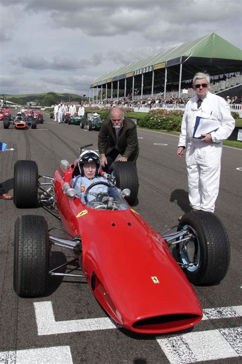 Two Men Standing Next To A Red Race Car On A Track With Other Cars In