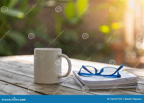 Close Up Of White Mug Cup Of Hot Coffee With Book And Reading Glasses On Wooden Table In Garden