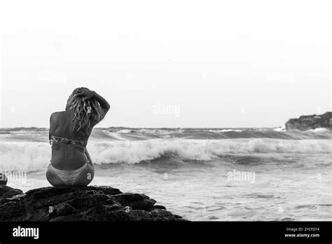 Blonde Woman With Loose Hair Dressed In A Bikini Sitting On A Cliff In Front Of The Sea Splashed