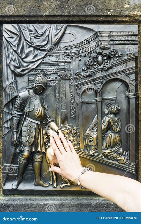 Tourist Rubs The Bronze Memorial Plaque Of Saint John Of Nepomuk On Charles Bridge In Prague For