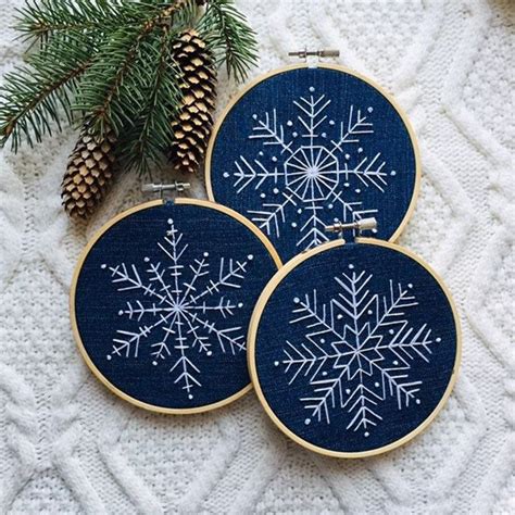 Two Cross Stitch Snowflakes Are On A White Blanket Next To Pine Cones