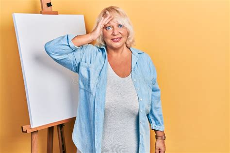 Middle Age Blonde Woman Standing By Painter Easel Stand Surprised With