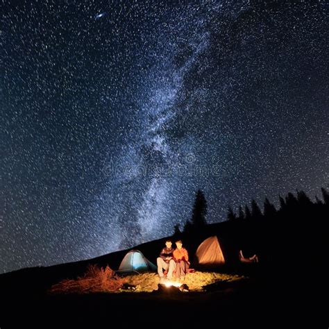 Couple Tourists Near Campfire And Tents Under Night Sky Full Of Stars