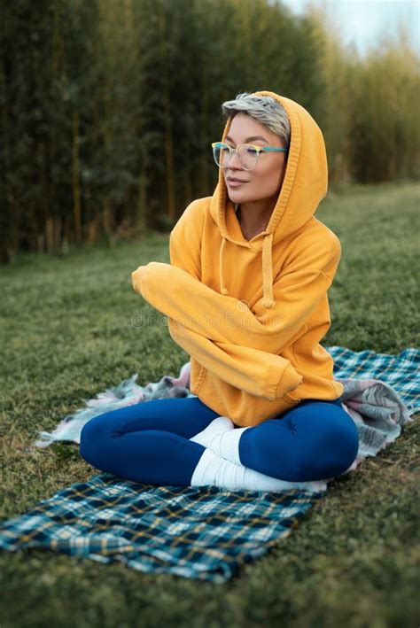 Cute Girl With Glasses In An Orange Hoodie Is Sitting In A Park In