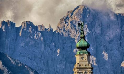 A Tall Building With A Clock On The Side Of It S Face In Front Of Mountains