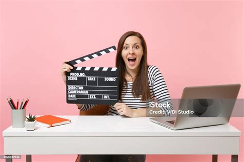 젊은 흥분 여자 잡아 고전적인 흑인 영화 Clapperboard 파스텔 핑크 배경에 고립 된 노트북으로 사무실에 앉아 있는 동안