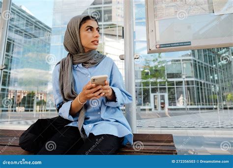 Young Muslim Woman Using Cellphone While Sitting On Bus Stop Stock