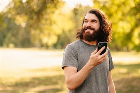 Cheerful Bearded Man With Long Hair Holding His Phone Is Looking Aside