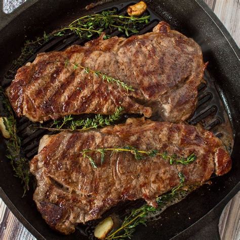 Two Steaks Cooking In A Cast Iron Skillet On A Wooden Table With Herbs