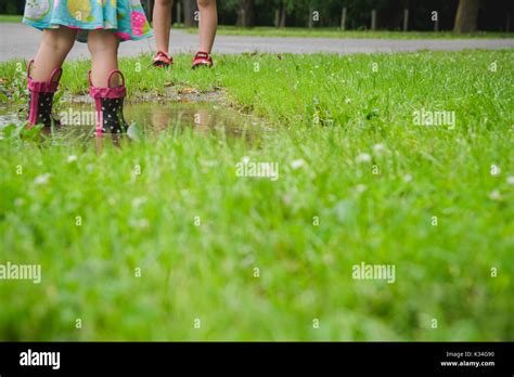 Children Stand In A Mud Puddle Wearing Rain Boots And With Feet Only