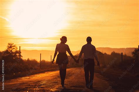 Stylish Couple Hold Each Other Hands While Walking Along The Roa Stock