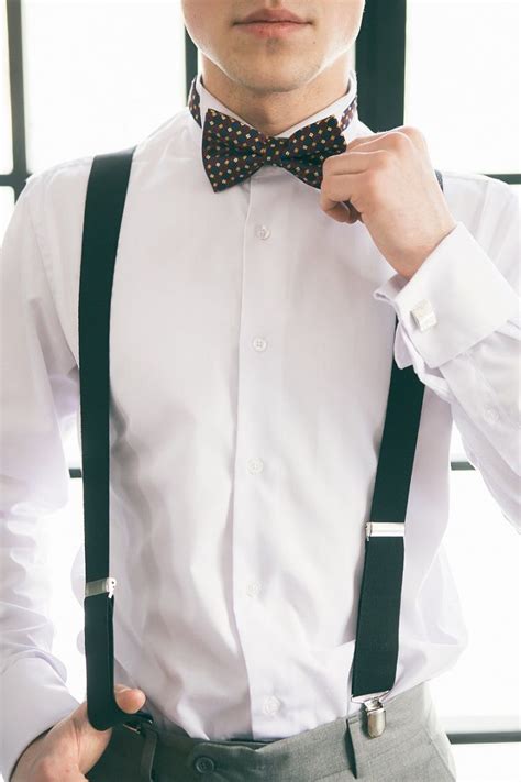 A Man In A White Shirt And Black Suspenders Is Adjusting His Bow Tie