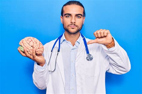 Young Handsome Man Wearing Doctor Coat Holding Brain With Angry Face