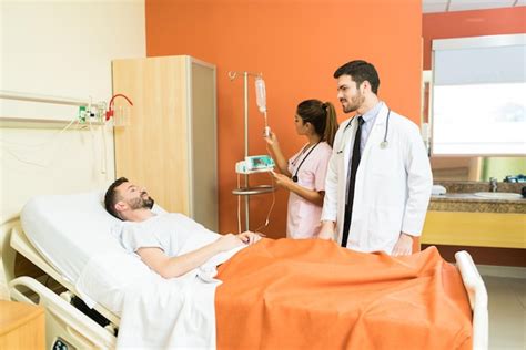 Male Doctor Talking To Patient Lying On Hospital Bed With Female