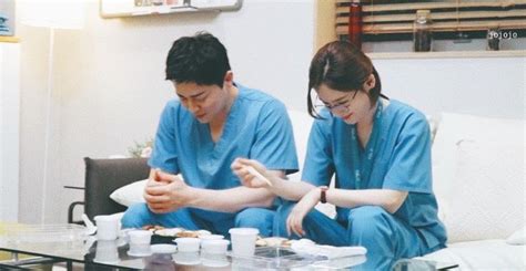 Two People In Scrubs Sitting On A Couch With Cups And Plates Of Food