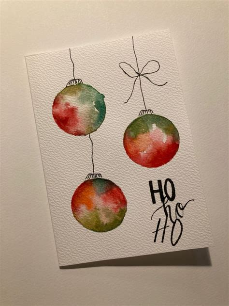 A Watercolor Christmas Card With Three Ornaments Hanging From It S