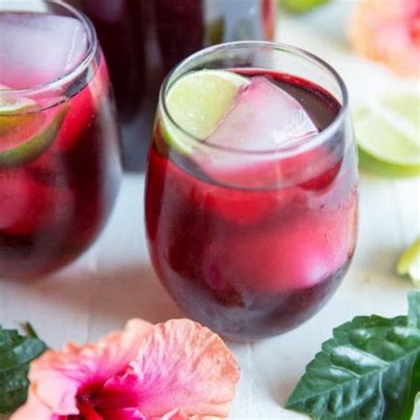 Two Glasses Filled With Red Liquid And Limes Next To Flowers On A White