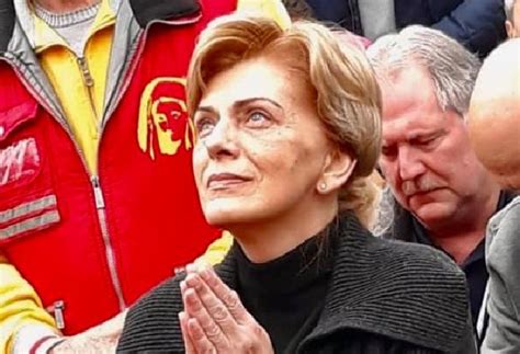 A Woman Standing In Front Of A Crowd With Her Hands Clasped To Her