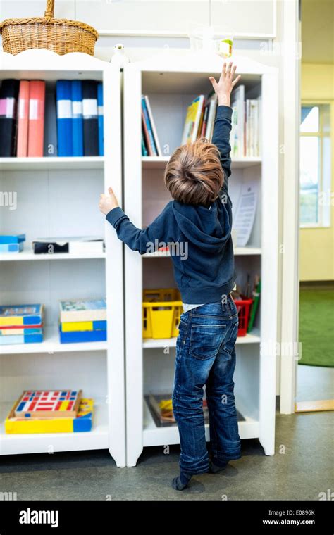 Rear View Of Boy Reaching For Container On Shelf In Kindergarten Stock