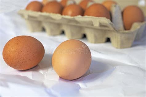 Two Eggs On A Table Near Brown Eggs In A Cardboard Container On A Light