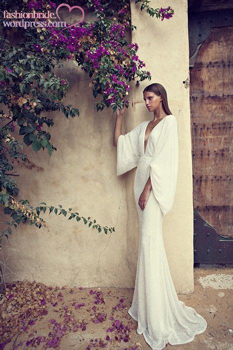 A Woman In A White Dress Standing Next To A Wall With Purple Flowers On It