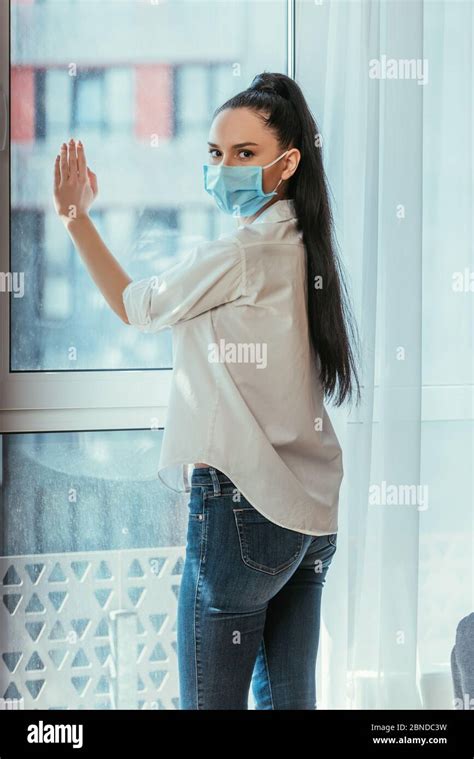 Sad Girl In Medical Mask Touching Window Glass And Looking At Camera