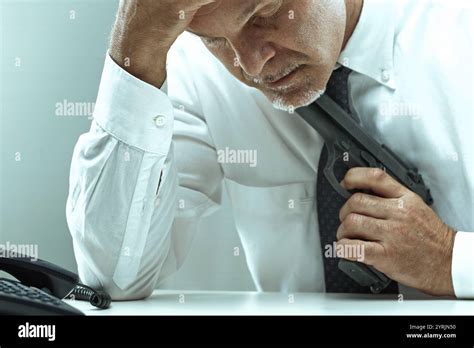 Stressed Mature Businessman Sitting At His Desk Gripping A Gun Under