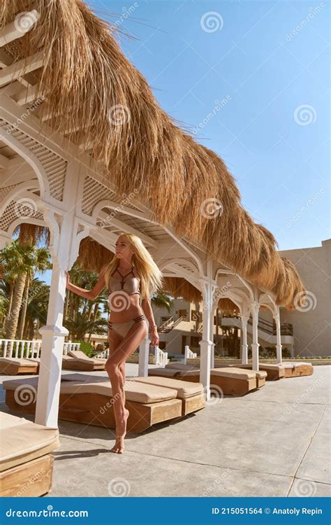 Woman Wearing Bikini Standing At Lounger Under Straw Canopy At Resort