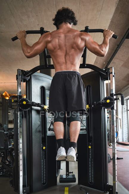 Back View Of Shirtless Man Performing Pull Ups On Exercise Machine While Training In Gym