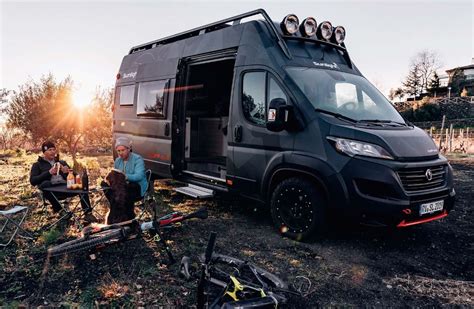 Two People Sitting In Front Of An Rv Parked On The Side Of A Dirt Road