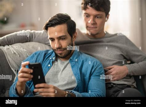 Cute Male Gay Couple Spend Time At Home They Are Lying Down On A Sofa And Use A Smartphone