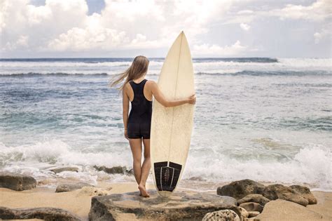 Rear View Of Woman Holding Surfboard While Standing On Rocky Shore At