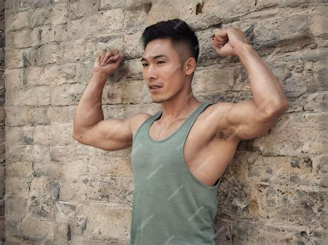 Portrait Of Muscular Asian Guy Flexing Biceps With Tank Top Leaning On Bricks Wall Premium Ai