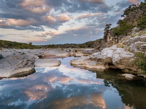 The Sky Is Reflecting In The Water Near Some Rocks And Trees On The