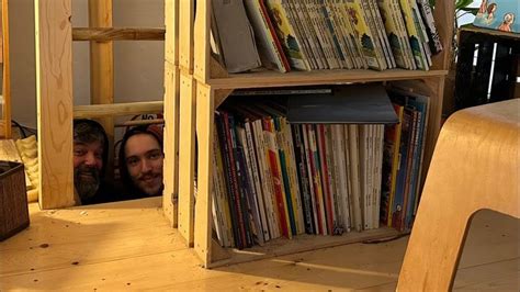 Two People Peeking Out From Behind A Bookshelf In A Room With Wooden Floors